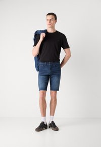 T-shirt nera, pantaloni corti di jeans blu e scarpe da ginnastica scure con calzini bianchi. Giacca di jeans appesa su una spalla, design minimale.