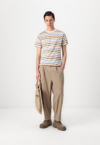 Striped cotton t-shirt with beige, green, orange, and blue hues; relaxed beige trousers; carrying a tan bag; textured footwear.