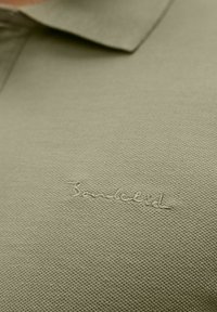 Oliven grøn polo-shirt med tekstureret stof og en krave. Har diskret broderet branding på venstre brystområde.