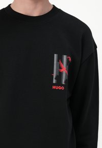 Felpa nera con collo a coste, caratterizzata da una grafica di un uccello rosso e la parola "HUGO" in bold rosso nell'area a sinistra del petto.