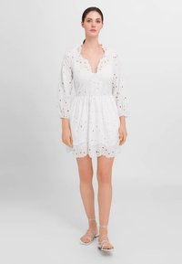 Rochie albă cu bust ajustat, decolteu în V și talie adunată. Prezintă un design brodat cu ochiuri și mâneci bufante. Margine ondulată la hem.