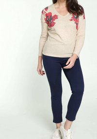 Beżowy sweter z długim rękawem z czerwonym kwiatowym wzorem na ramionach, w połączeniu z granatowymi spodniami slim-fit i białymi sneakersami.