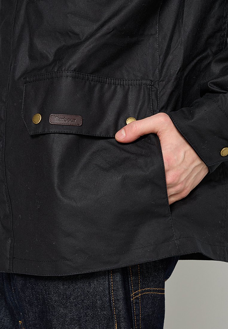 Veste noire imperméable avec une finition texturée, dotée d'une poche avant avec une étiquette marron et des boutons-pression dorés, portée sur un jean.