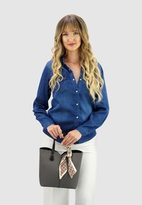 Grijze boodschappentas met een gestructureerde afwerking, zwarte handvatten en een crèmekleurige sjaal met patroon die om het handvat is geknoopt. Het model draagt een blauwe spijkerbloes en witte broek.