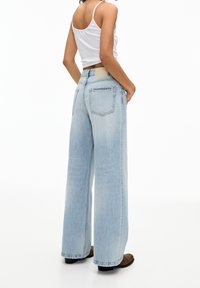 Jean large taille haute de couleur bleu clair avec poches arrière et texture lisse. Porté avec un débardeur blanc et des bottes marron.