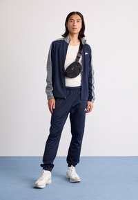 Navy tracksuit with gray accents, zip jacket, elastic waistband pants, paired with a black waist bag and white sneakers on a blue background.