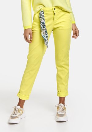 Personne portant un pantalon jaune vif retroussé avec un foulard fleuri dans la poche, un pull jaune et des baskets blanches et beiges.