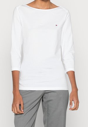 Personne portant une chemise blanche à manches longues avec un petit logo sur la poitrine et un pantalon gris, debout devant un fond uni.
