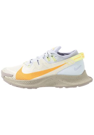 Chaussure de sport beige clair et grise avec un swoosh orange, une sangle jaune au talon, une fermeture lacée et une semelle texturée, vue de côté.