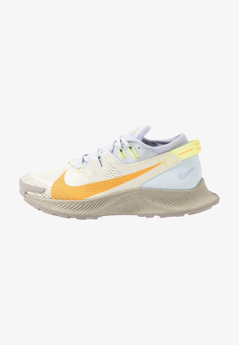 Chaussure de sport beige clair et grise avec un swoosh orange, une sangle jaune au talon, une fermeture lacée et une semelle texturée, vue de côté.