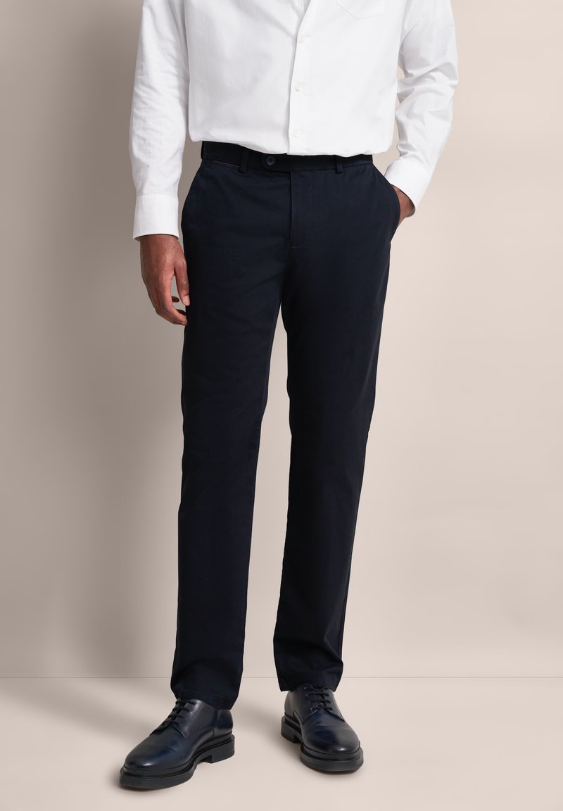 Mężczyzna w czarnych eleganckich spodniach, czarnych skórzanych butach i białej koszuli z guzikiem, z jedną ręką w kieszeni na beżowym tle.