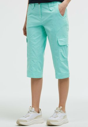 Hell türkise knielange Cargo-Shorts, getragen von einer stehenden Person, kombiniert mit weißen und beigen Sneakers vor einem einfarbigen Hintergrund.
