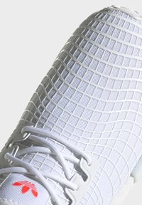 Zapatilla deportiva blanca con un upper texturizado en forma de cuadrícula, cordones blancos y un pequeño acento de logo rojo cerca de la base.