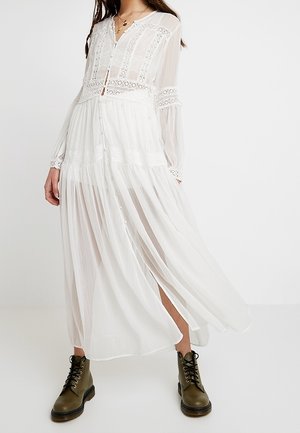 Femme portant une robe maxi blanche à manches longues, boutonnée et ornée de dentelle, associée à des bottines de combat à lacets vert olive.