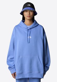 Haut de hoodie bleu clair avec poche avant et capuche à cordon, arborant un logo blanc sur la poitrine ; associé à un chapeau seau noir avec des accents bleus.