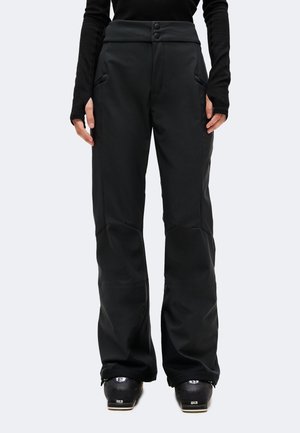 Personne portant un pantalon de neige noir et des bottes de ski noires, debout face à l’avant avec les mains détendues sur les côtés.