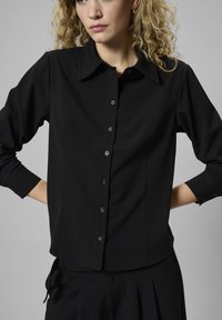 Melnā krekla ar pogām ar apkakli, garām piedurknēm un spīdīgām pogām. Gluda auduma ar pielāgotu piegriezumu, ar sānu šķēlumiem apakšmalā.