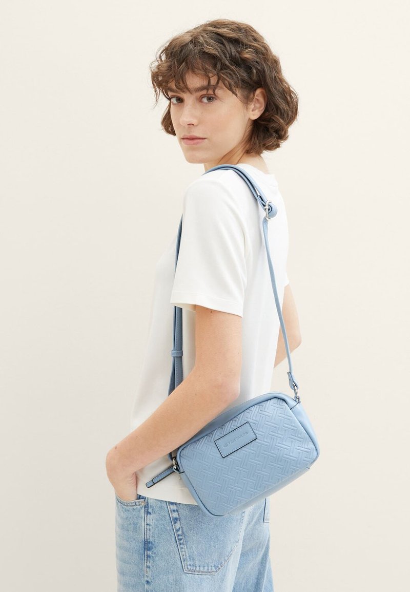 Hellblaues Crossbody-Bag mit strukturiertem Muster, rechteckiger Form und verstellbarem Träger. Glattes Material mit einem kleinen Logo-Patch auf der Vorderseite.