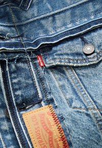Denimstof in lichte blauwe tint. Kenmerkt zich door een rode Levi's-label, metalen knoop en contrasterende stiksels. Zichtbare textuurpatronen en merketiket.