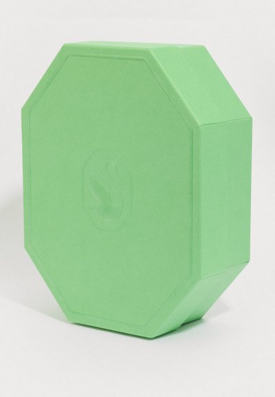Block de yoga octogonal vert avec un subtil logo de cygne en relief sur une face, posé à la verticale sur une surface blanche.