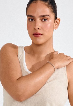 Kobieta o brązowych włosach w beżowym topie dotyka swojego ramienia, nosząc cienką srebrną bransoletkę, na jednolitym białym tle.