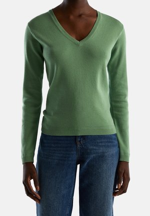 Person iført en grøn langærmet V-hals sweater og blå jeans, stående mod en ensfarvet lys baggrund.