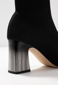 Černé látkové kotníkové boty s robustním lesklým černým a šedým gradientním podpatkem, který má vroubkovanou strukturu a elegantní design.