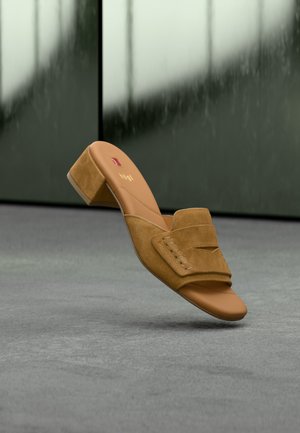 Ταμπά σουέτ mule με ανοιχτό δάχτυλο και χαμηλό χοντρό τακούνι, που αιωρείται πάνω από γκρι επιφάνεια, ορατή η επωνυμοποιημένη εσωτερική σόλα.