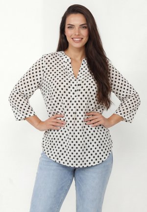 Femme aux longs cheveux bruns souriante, portant un chemisier blanc à pois noirs et un jean bleu clair, debout les mains sur les hanches.