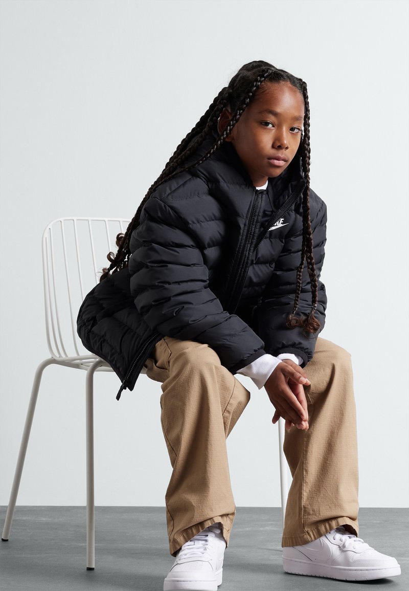 Svart puffjacka med hög krage och hel dragkedja, bärs över en vit skjorta. Beiga vida byxor och vita sneakers fulländar outfiten.