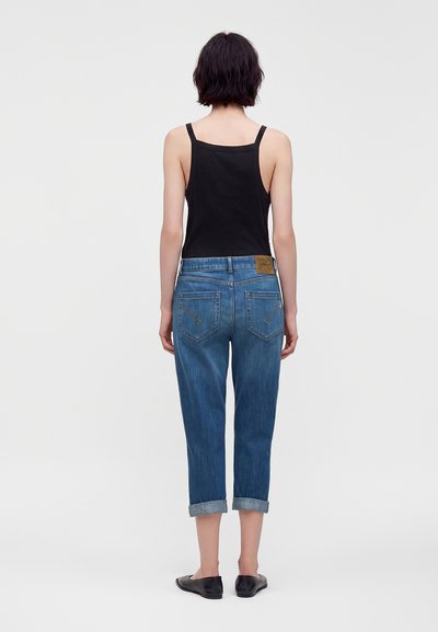 Personne aux cheveux courts et foncés porte un débardeur noir, un jean bleu retroussé et des chaussures noires sans lacets, debout de dos sur fond blanc.