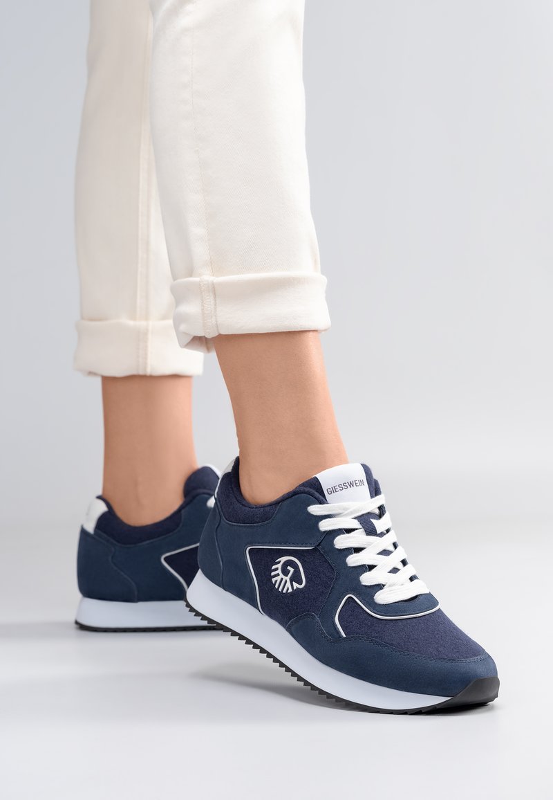 Voeten met marineblauwe Giesswein sneakers met witte veters en opgerolde, crèmewitte broekspijpen op een lichte achtergrond.