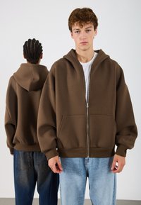 Rjava zip-up kapuca iz mehkega materiala, z prostornim krojem, sprednjimi žepi in velikim kapuco, v kombinaciji z svetlo modrimi jeans hlačami.