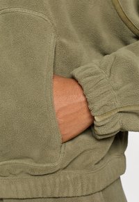 Sudadera de felpa en verde oliva, con un bolsillo tipo canguro, puños acanalados y una tela suave y texturizada. Destacada con un ribete en contraste.