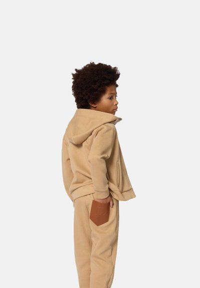 Tan hoodie met een voorzak, gecombineerd met bijpassende joggerbroek met een bruine leren patch op de achterzak.