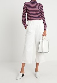 Maglione a collo alto a righe blu scuro con strisce rosa, abbinato a pantaloni bianchi a gamba larga. Mule bianche e una piccola borsa strutturata con dettagli neri.