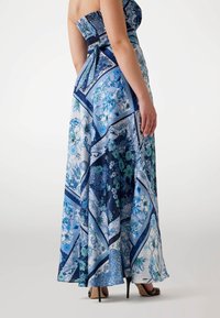Robe maxi à imprimé floral bleu et blanc, avec un bodice smocké élastiqué, une jupe fluide et un dos noué. Chaussures à talons hauts visibles.