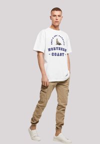 Weißes übergroßes T-Shirt mit einem Seehund-Graphik und blauer Schrift. Sandfarbene Cargo-Hosen mit mehreren Taschen und elastischen Bündchen. Weiße Sneakers.