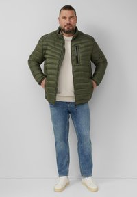 Grüne, isolierte Steppjacke mit horizontalen Nähten, hohem Kragen, Reißverschluss und seitlichen Taschen, getragen über einem beigen Pullover und blauen Jeans.