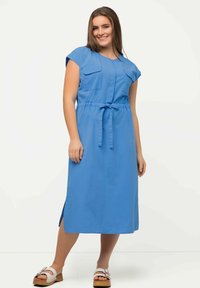Blaues Kleid mit kurzen Ärmeln, zwei Fronttaschen, tailliert und mit Bindeband. Glatter Stoff, knöchellang, mit Schlitzen an den Seiten. Getragen mit weißen Sandalen.