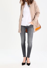 Chaqueta bomber beige claro con un forro interior naranja, combinada con una camisa blanca de botones y unos jeans grises ajustados. Tacones altos negros completan el conjunto.