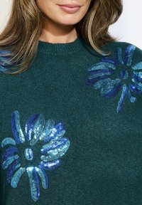 Maglione verde lavorato a maglia con motivi floreali blu in paillettes sul petto. Collo rotondo e texture morbida. Inquadratura ravvicinata.