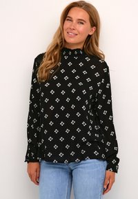Zwarte blouse met bloemachtige ruitpatronen in wit en groen. Hoge halslijn met een gerimpelde rand, lange mouwen en een losse pasvorm.