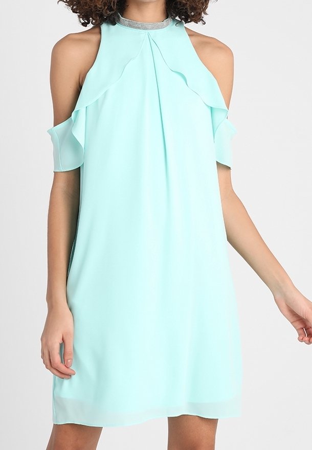 Femme portant une robe bleu clair sans manches avec des découpes aux épaules et des détails à volants, debout devant un fond uni.
