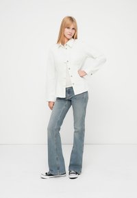 Valge teksajakk nööpkinnitusega ja kahe rinnataskuga, kombineeritud helesiniste lahenevate teksapükstega ja mustade tossudega. Ühtlane taust.