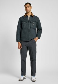 Mørkeblå jakke med brun krave, to brystlommer, lynlås; parret med grå cargo bukser og sort-hvide sneakers.
