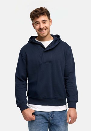 Lächelnder junger Mann, der einen dunkelblauen Hoodie über einem weißen T-Shirt und blauen Jeans trägt, vor einem einfarbigen hellen Hintergrund stehend.