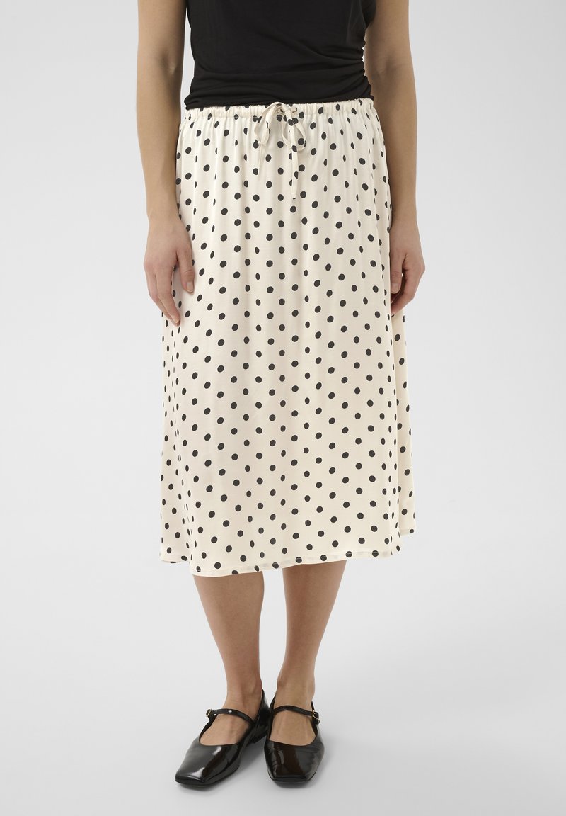 Femme portant une jupe midi blanche à pois noirs, un haut noir et des chaussures Mary Jane noires, debout sur un fond clair uni.