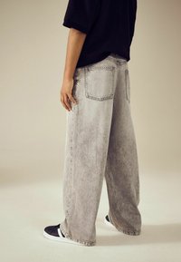 Hellgraue weite Jeans mit einem verblassten Muster, ausgestattet mit zwei Gesäßtaschen und umgeschlagenen Säumen, kombiniert mit schwarzen Slippers.