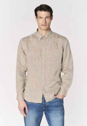 Uomo con camicia beige a maniche lunghe abbottonata e jeans blu, in piedi con una mano in tasca davanti a uno sfondo uniforme.